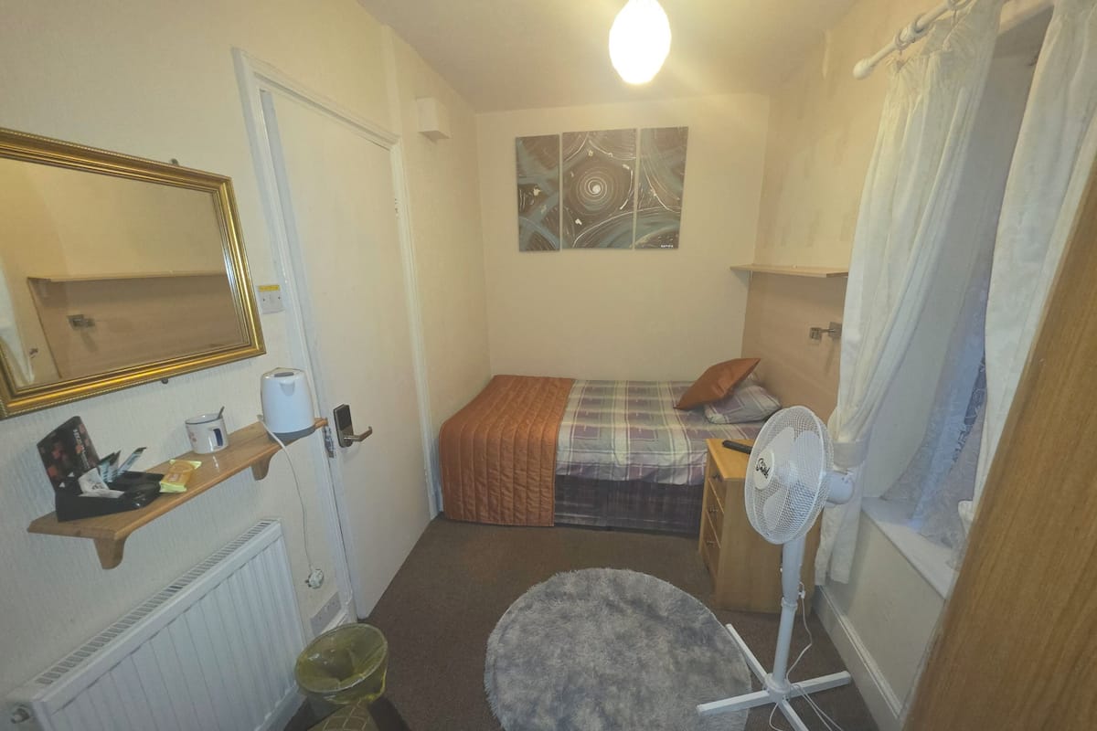 A small, compact room with a bed, a desk, a chair, a radiator, a mirror, a fan, and a window with curtains.