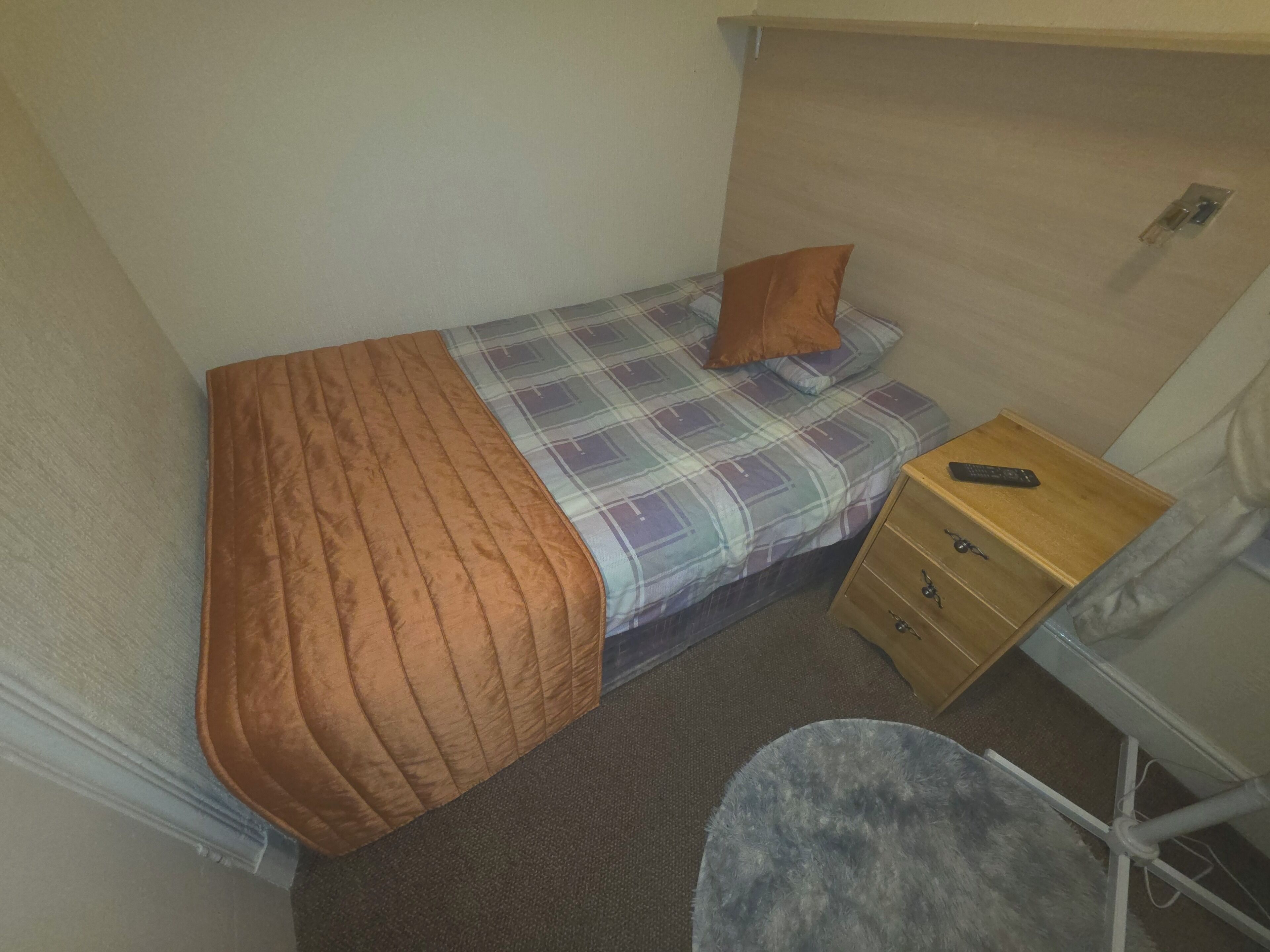 A small, single-bed room with a plaid bedspread, a brown blanket, a wooden nightstand with a remote control, and a small chair.