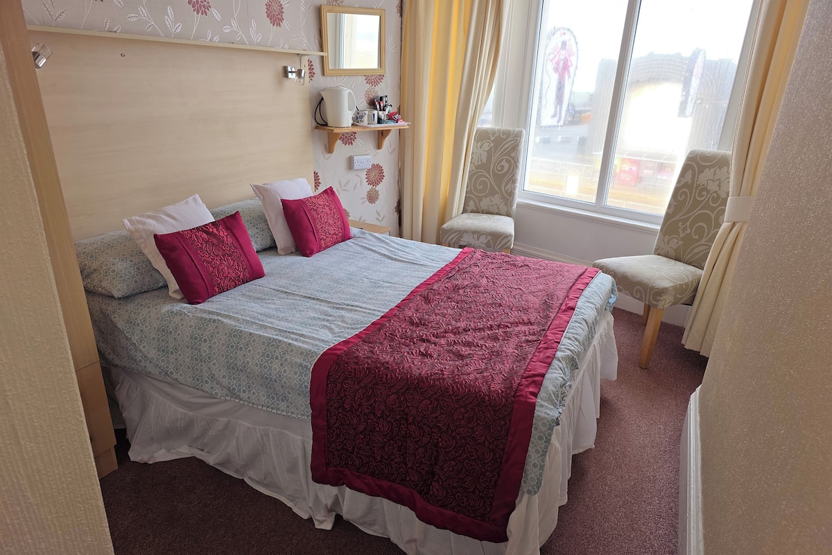 A neatly made bed with a patterned bedspread and pillows, a bedside table with a kettle and personal items, a chair with a floral pattern, and a window with a view of the outside.