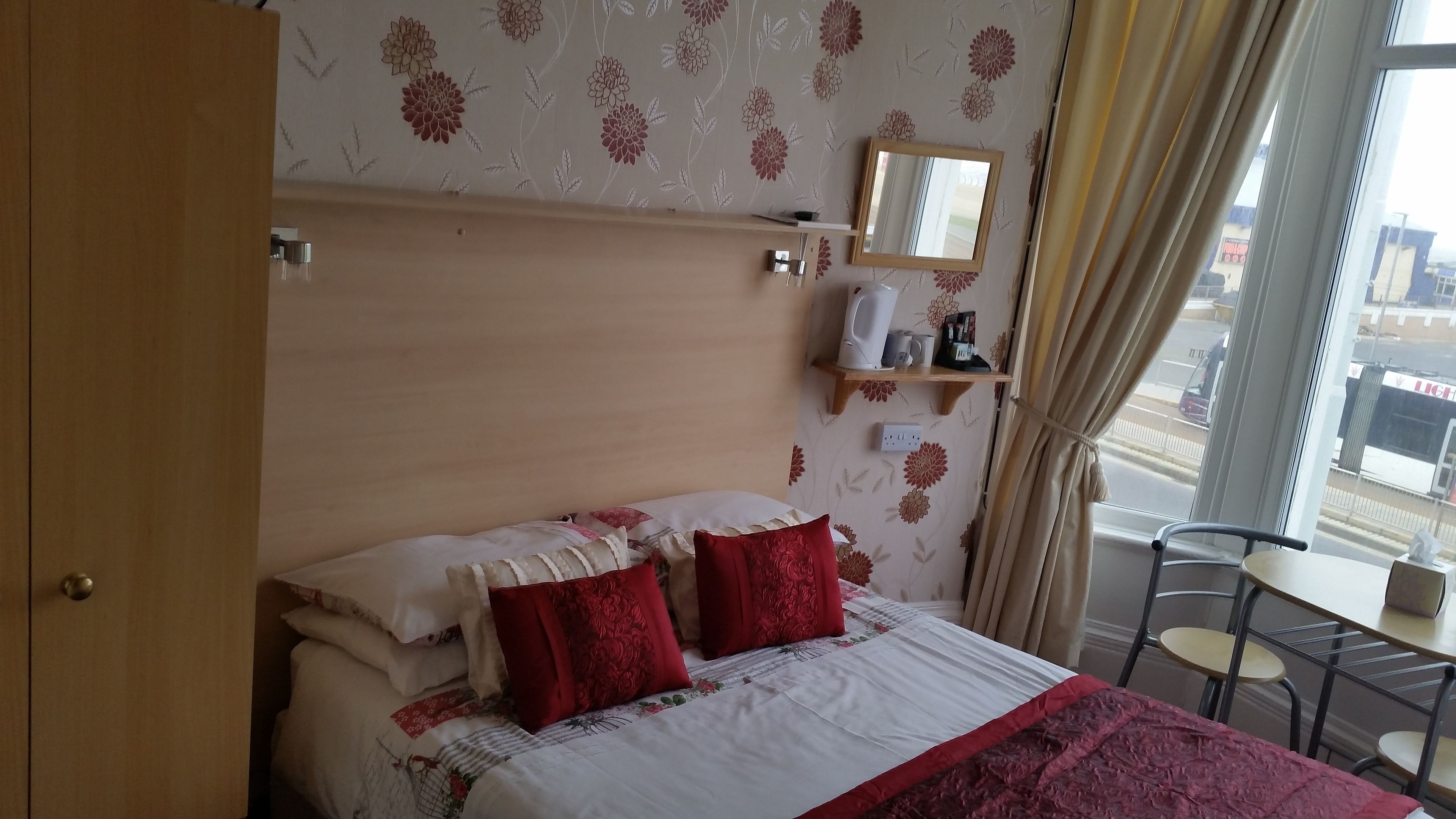 A hotel room with a bed, a bedside table, a chair, a window with curtains, and a wall-mounted shelf with a mirror and a kettle.