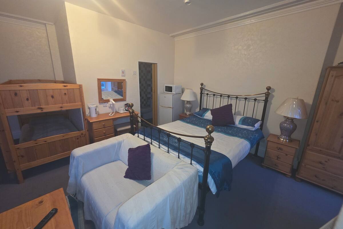 A bedroom with two single beds, a wooden bunk bed, a desk with a telephone, a mirror, a bedside table with a lamp, and a wardrobe.