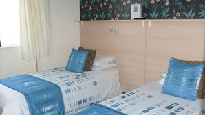 A room with two beds, each with a blue and white patterned bedspread. There is a wooden wardrobe above the beds with a floral wallpaper design. - The Sands Hotel (Blackpool)