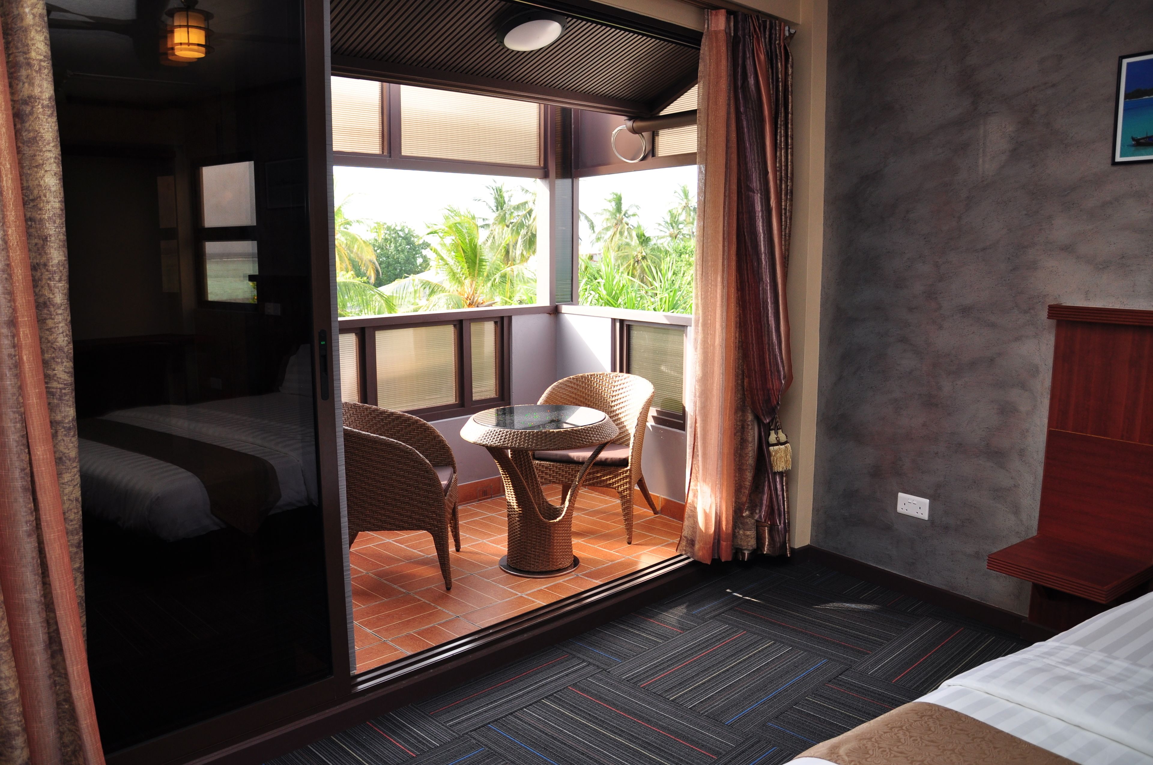 Habitación de hotel con cama, una pequeña mesa con sillas, vistas a palmeras y un balcón con cortinas.