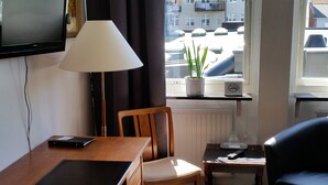 Una habitación con escritorio, silla, lámpara y televisor en la pared. Se ve una ventana con vistas a edificios.