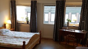 Un dormitorio con cama, escritorio y silla. Hay cortinas en las ventanas, una lámpara en el escritorio y un radiador en la pared.