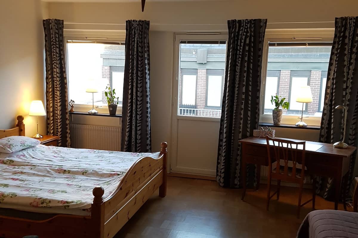 A bedroom with a bed, a desk, and a chair. There are curtains on the windows, a lamp on the desk, and a radiator on the wall.