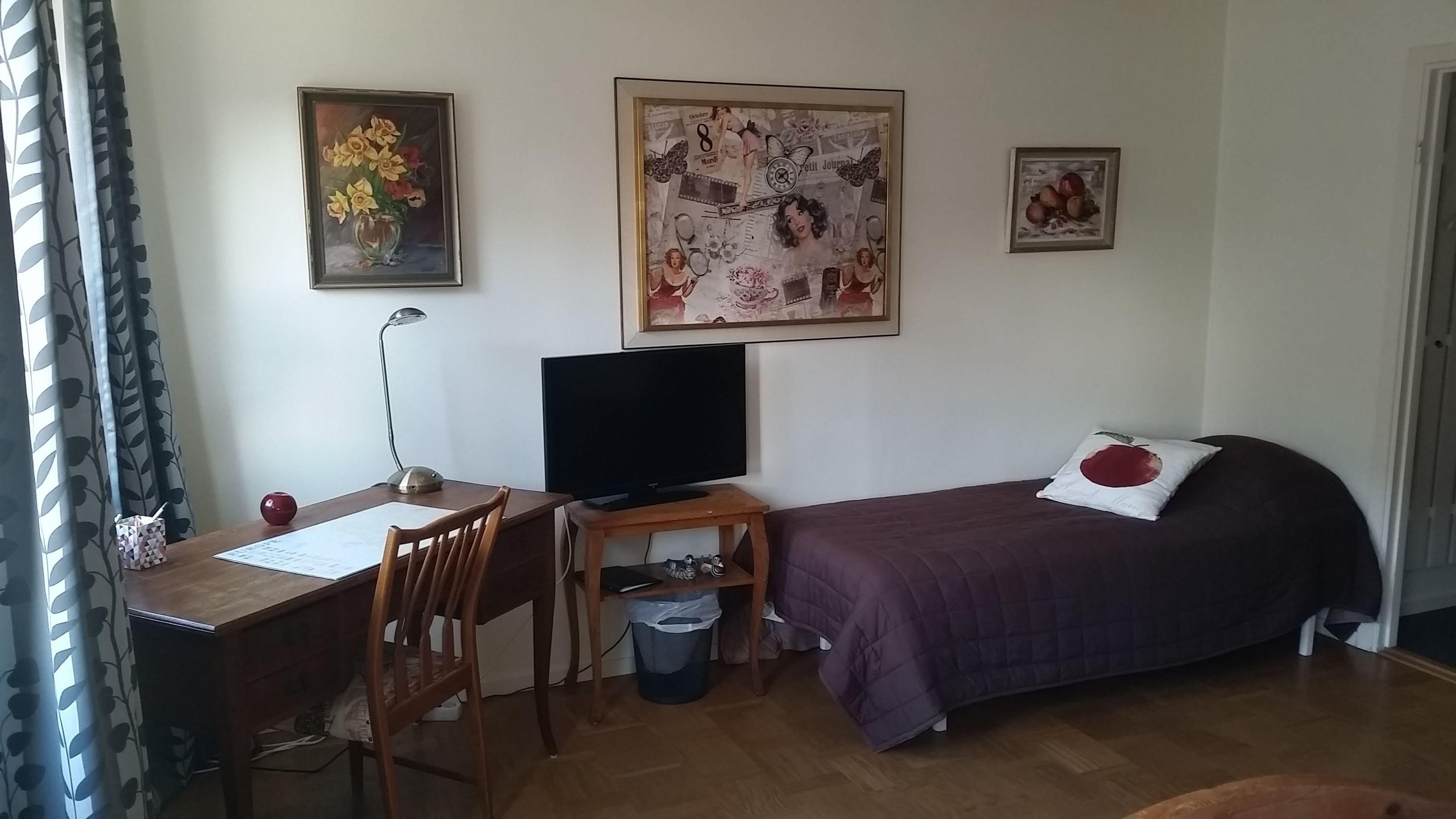 A room with a wooden desk, a chair, a television, a bed with a purple bedspread, and two framed pictures on the wall.