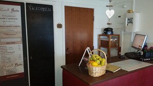 Un mostrador de recepción de hotel con una pizarra que muestra las tarifas de las habitaciones, una computadora y una cesta de fruta.