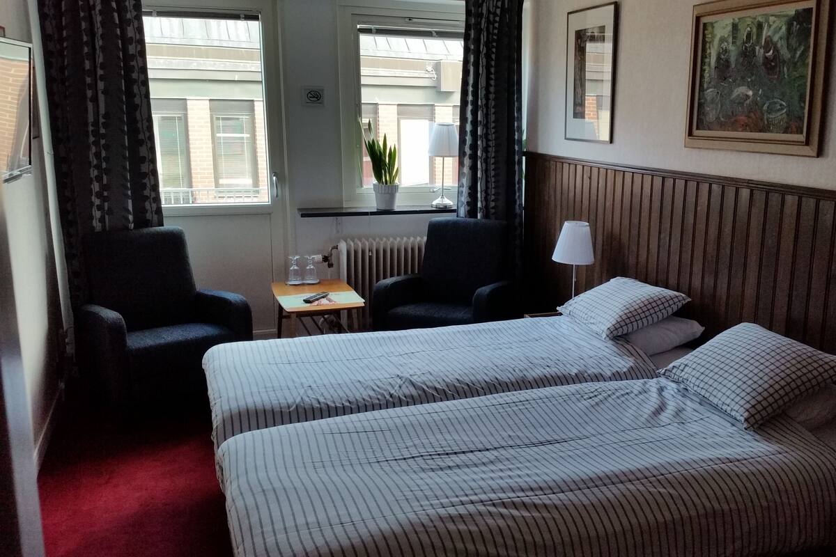 A hotel room with two beds, a desk, a chair, a radiator, and a window with curtains.