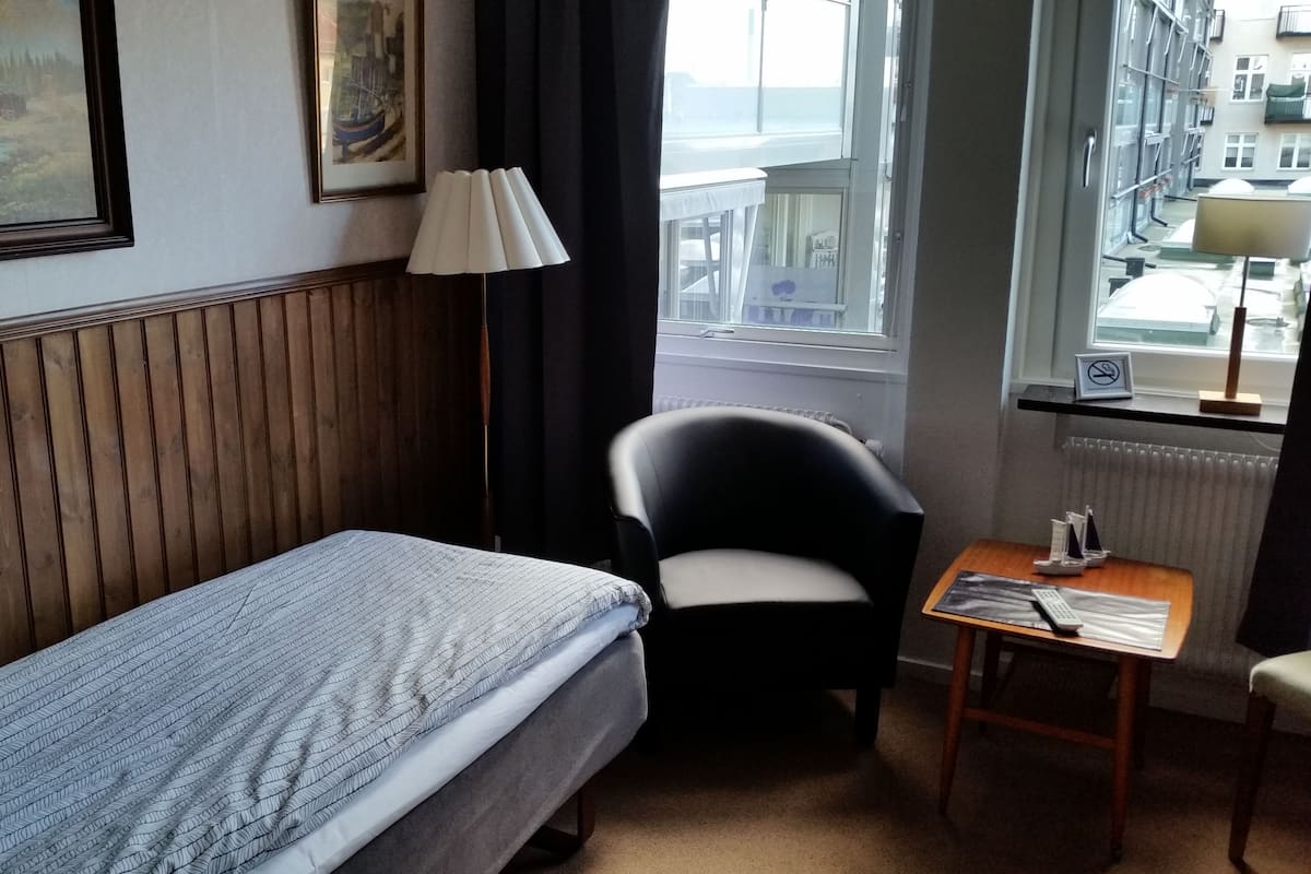 A hotel room with a single bed, a chair, a small table, a lamp, and a window with a view of buildings.