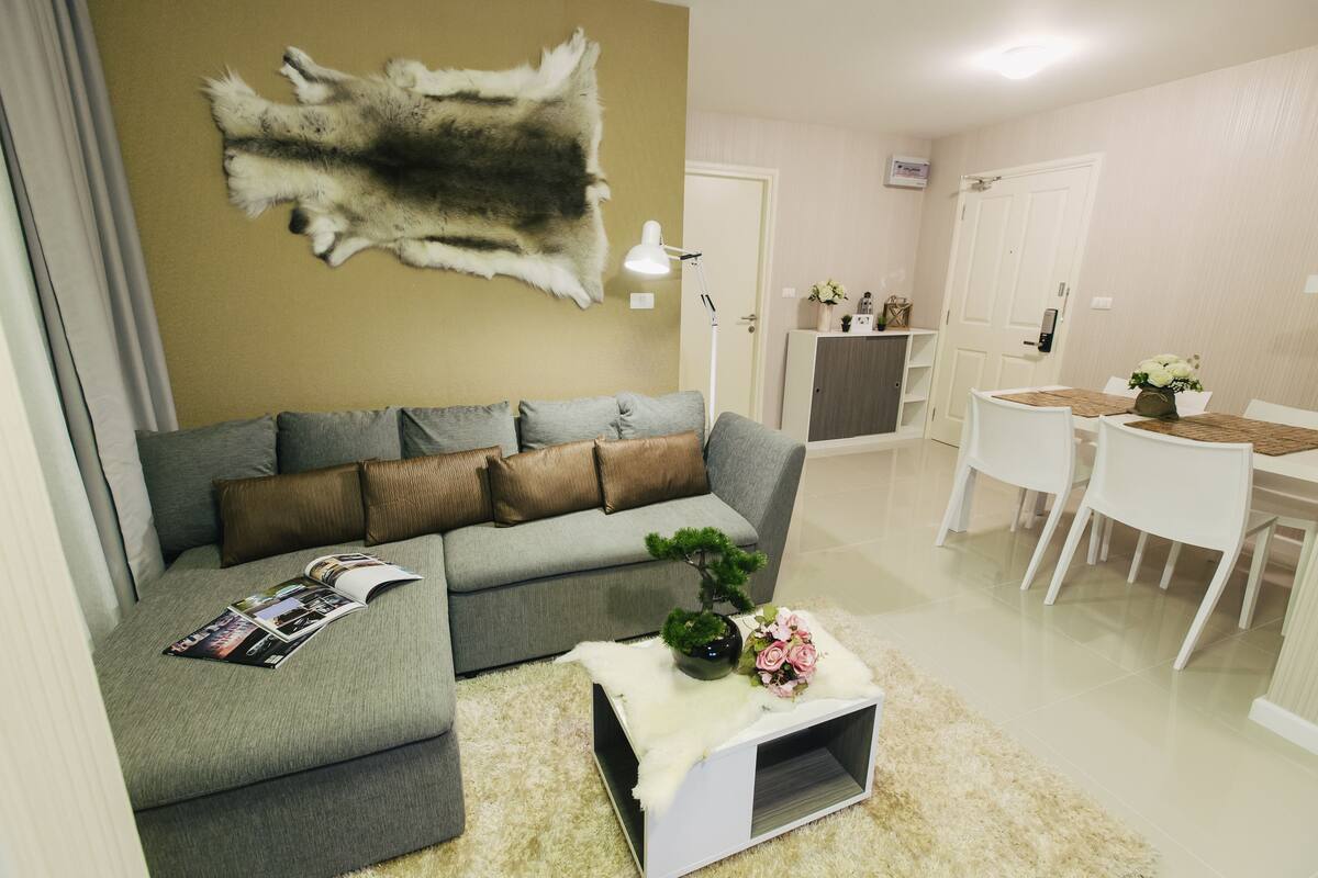 A living room with a grey sectional sofa, a white coffee table with a magazine, a small plant, and a rug. There is a dining area with a white table and chairs, and a door in the background.