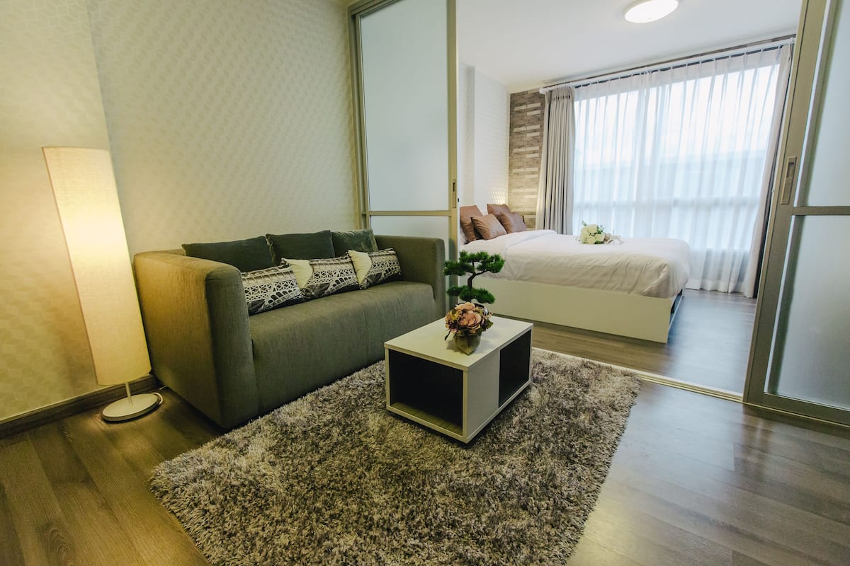 A modern hotel room with a sofa, a small side table with a plant, a bed with a white duvet, and a large window with curtains.