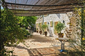 An outdoor patio with stone flooring, metal chairs, potted plants, and a stone building with shutters. - La Ferme d'Augustin (Ramatuelle)