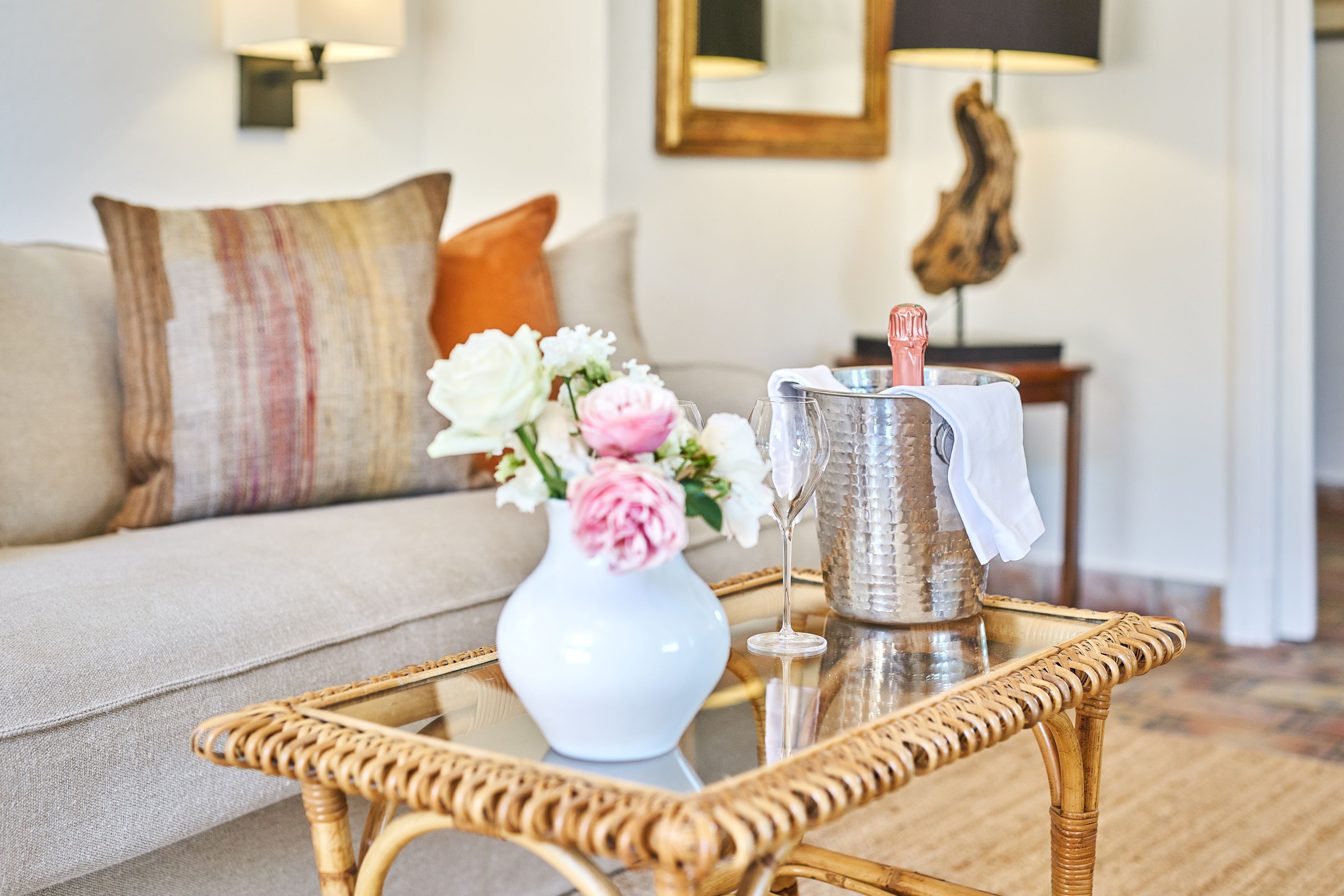 A wicker table with a white vase and a silver container, a beige sofa with a striped pillow, and a wall-mounted lamp.