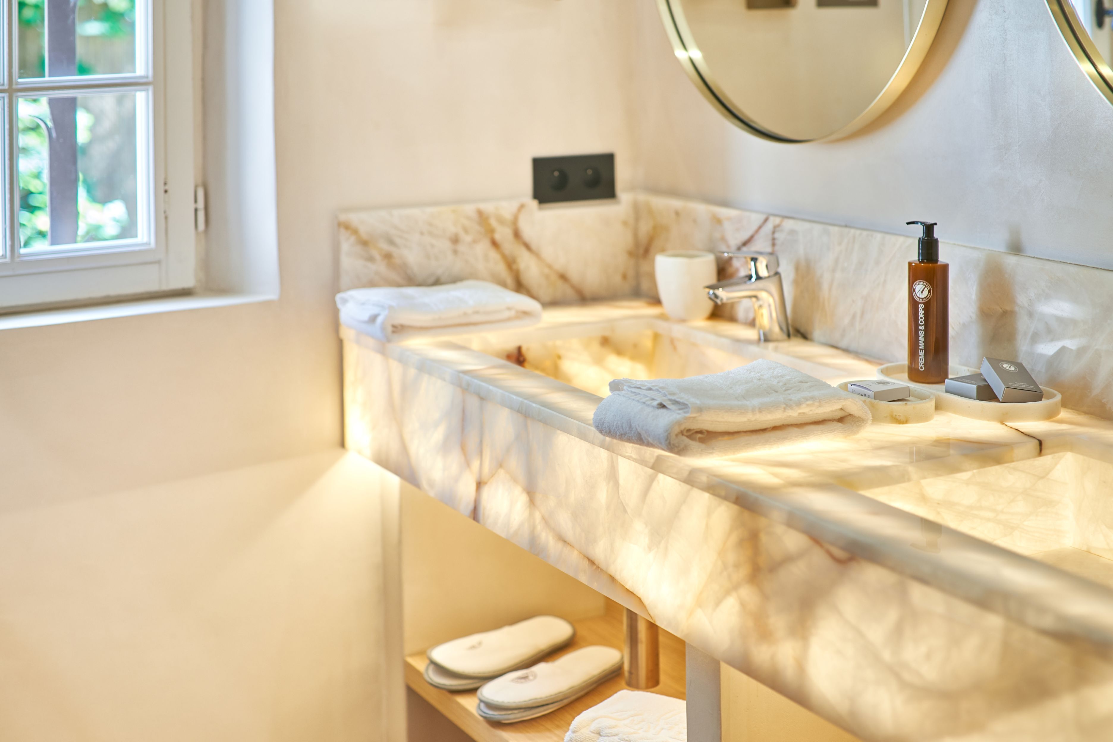 Une salle de bain avec un comptoir en marbre, un lavabo, un miroir, une bouteille de shampoing et une paire de pantoufles.