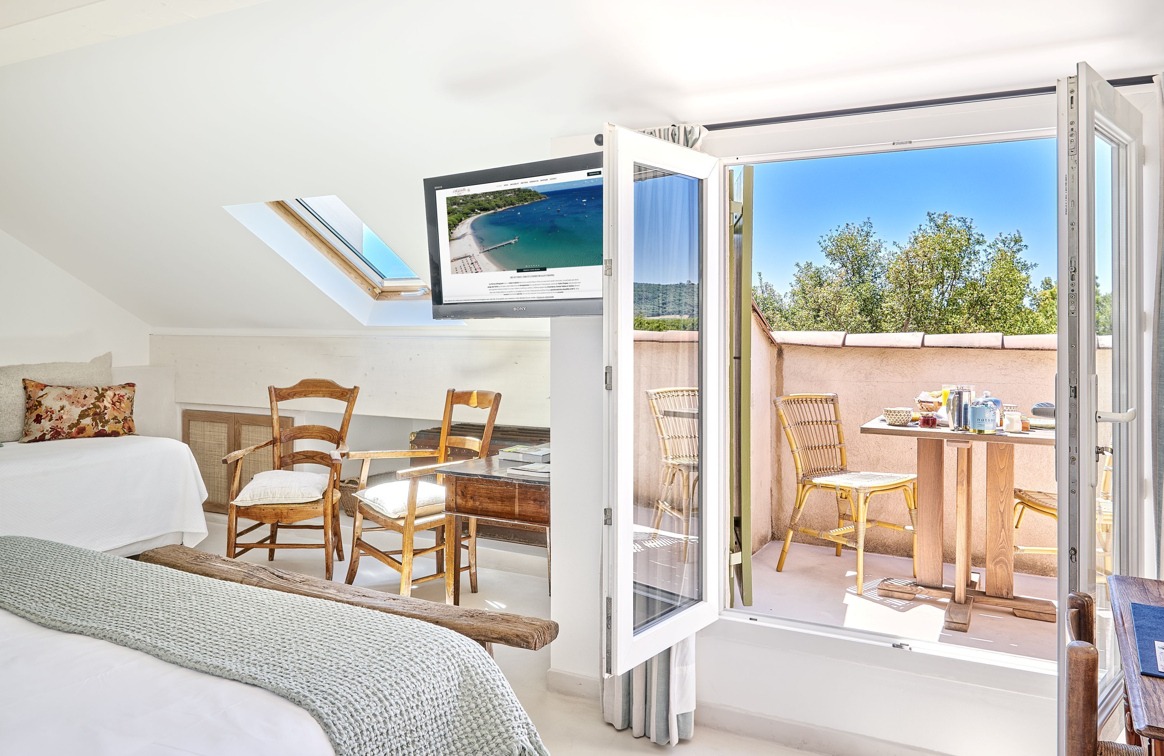 Ein Schlafzimmer mit einem Bett, einem Schreibtisch und einem Sessel, sowie ein Balkon mit Tisch und Stühlen und einem Fernseher, der einen Strand zeigt.