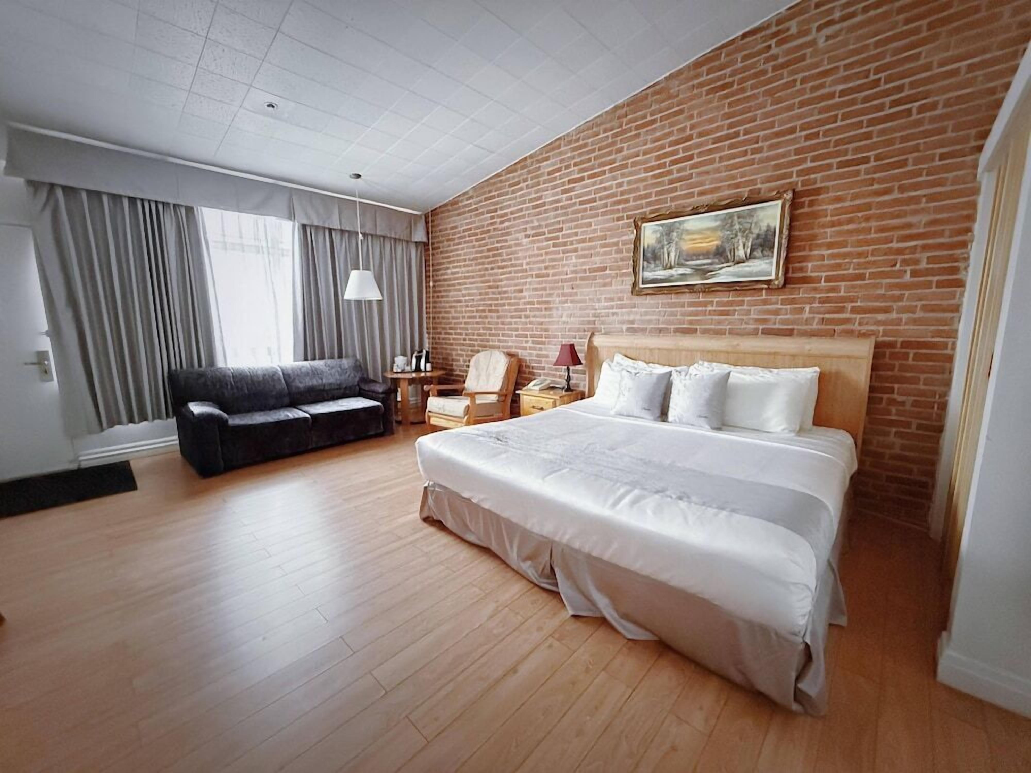 Ein Hotelzimmer mit einem großen Bett, einer Couch, einem Schreibtisch mit Lampe und einem Gemälde an der Backsteinwand.