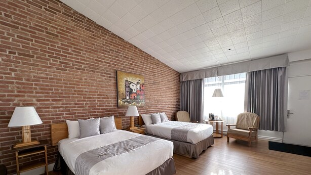 A hotel room with two beds, a brick wall, a painting, a window with curtains, a chair, and a desk with a lamp. - Hotel La Caravelle (Baie Comeau)