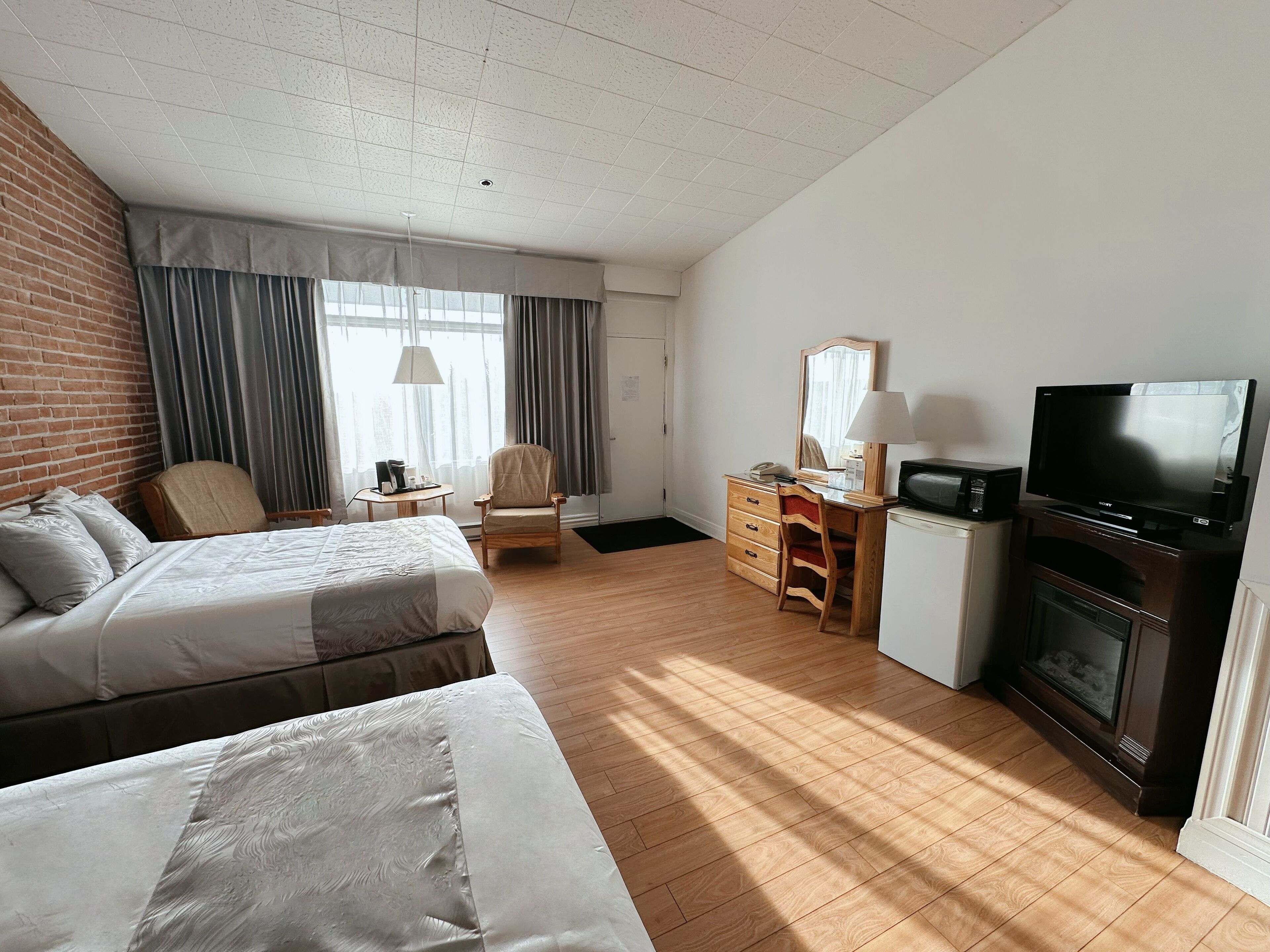 A hotel room with two beds, a desk with a TV, a fireplace, and a large window with curtains.