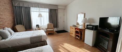 A hotel room with two beds, a desk with a TV, a fireplace, and a large window with curtains.