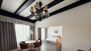 A hotel room with a bed, a dining table with chairs, a chandelier, and large windows with curtains. - Hotel La Caravelle (Baie Comeau)