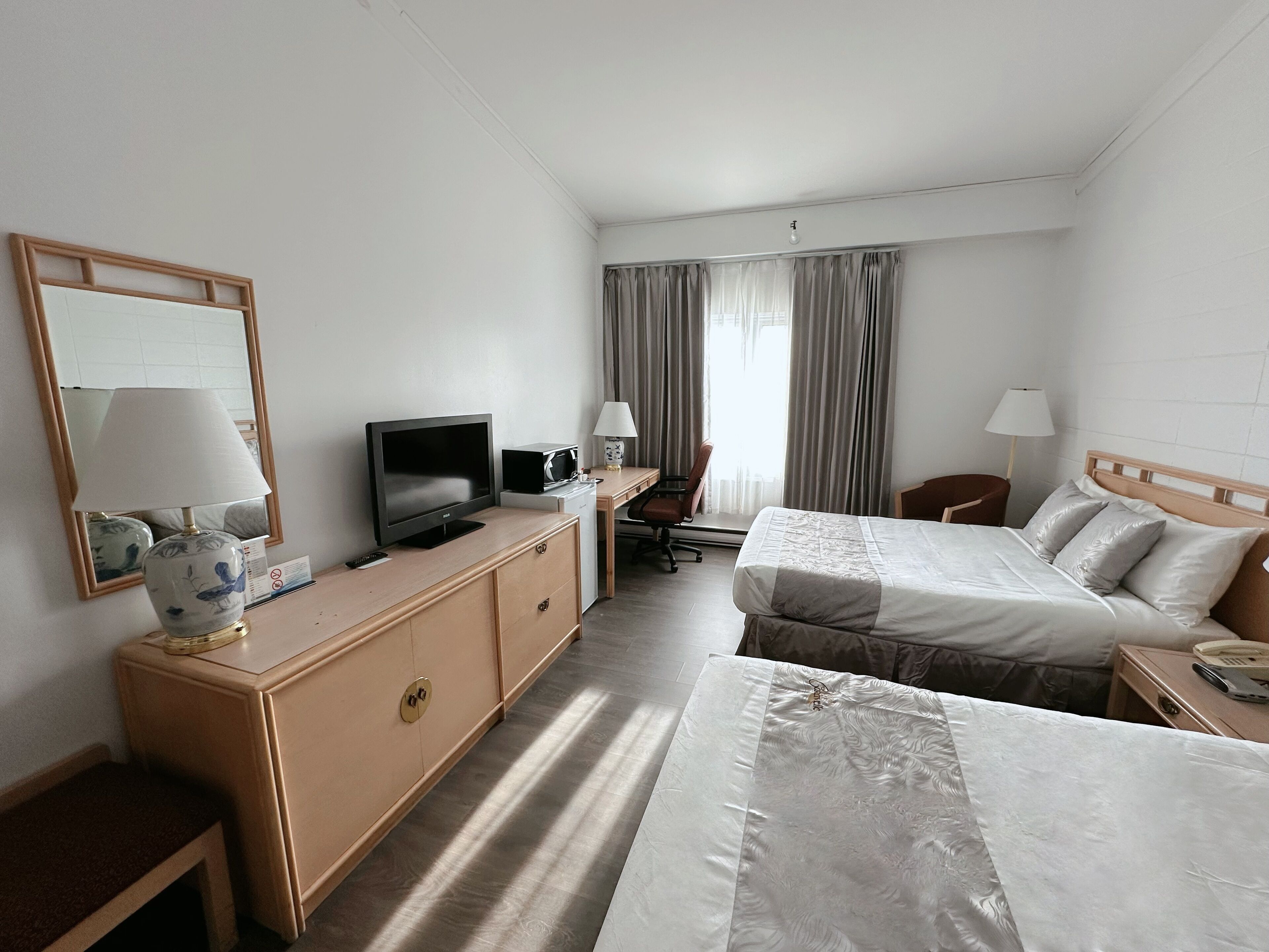 Habitación de hotel con una cama, un escritorio con televisor, una silla, una lámpara y una ventana con cortinas.