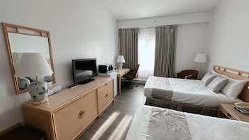 Habitación de hotel con una cama, un escritorio con televisor, una silla, una lámpara y una ventana con cortinas.