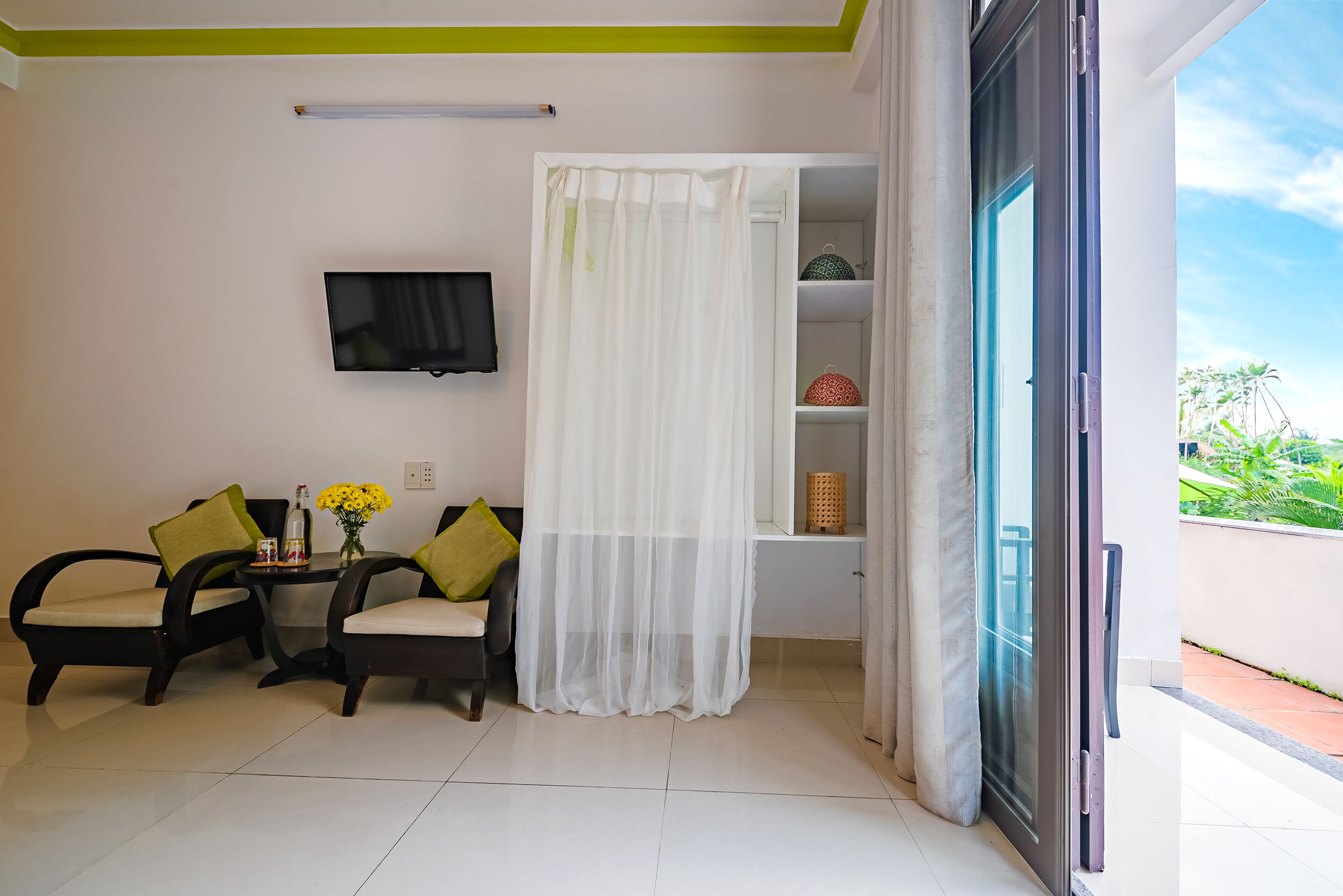A room with a TV, two chairs, a table with a vase of flowers, a white curtain, and a sliding glass door to an outdoor area.