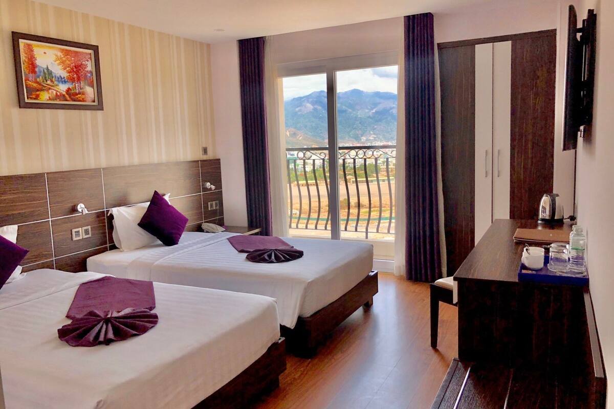 Une chambre d’hôtel avec deux lits, un bureau, un tableau au mur et un balcon offrant une vue sur la montagne.