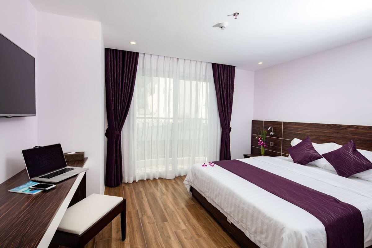 Une chambre d’hôtel moderne avec un bureau en bois, un ordinateur portable et un lit avec une literie blanche et violette.