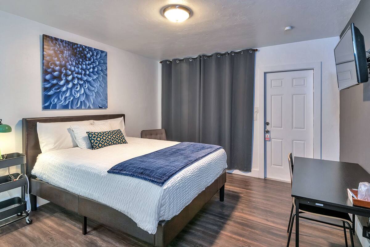 A bedroom with a bed, a desk, a chair, a painting on the wall, and a door.