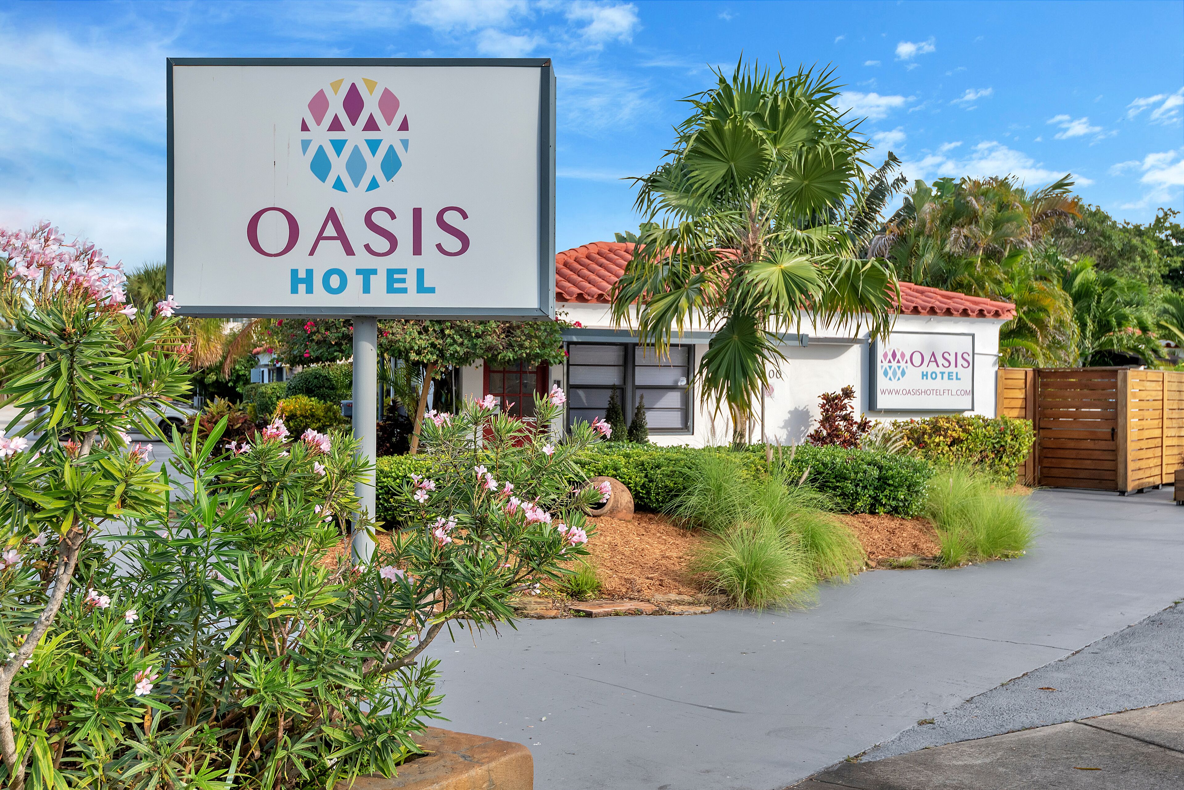 Placa do Hotel Oasis com um logotipo, cercada por vegetação e um edifício com telhado de telhas vermelhas.