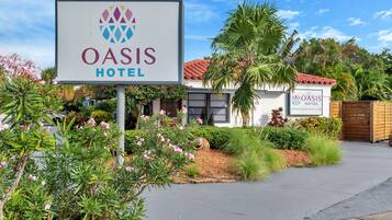 Placa do Hotel Oasis com um logotipo, cercada por vegetação e um edifício com telhado de telhas vermelhas.