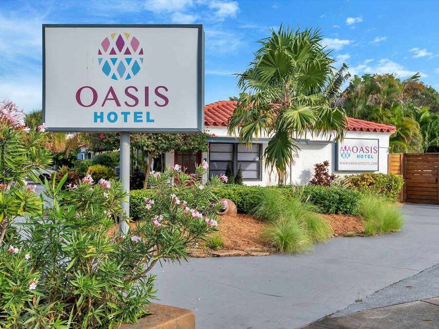 Placa do Hotel Oasis com um logotipo, cercada por vegetação e um edifício com telhado de telhas vermelhas.