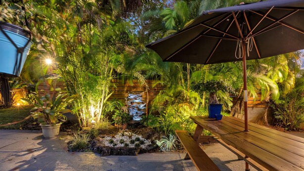 A patio area with a wooden bench, an umbrella, and a small waterfall feature surrounded by lush greenery. - Oasis Hotel (Fort Lauderdale)
