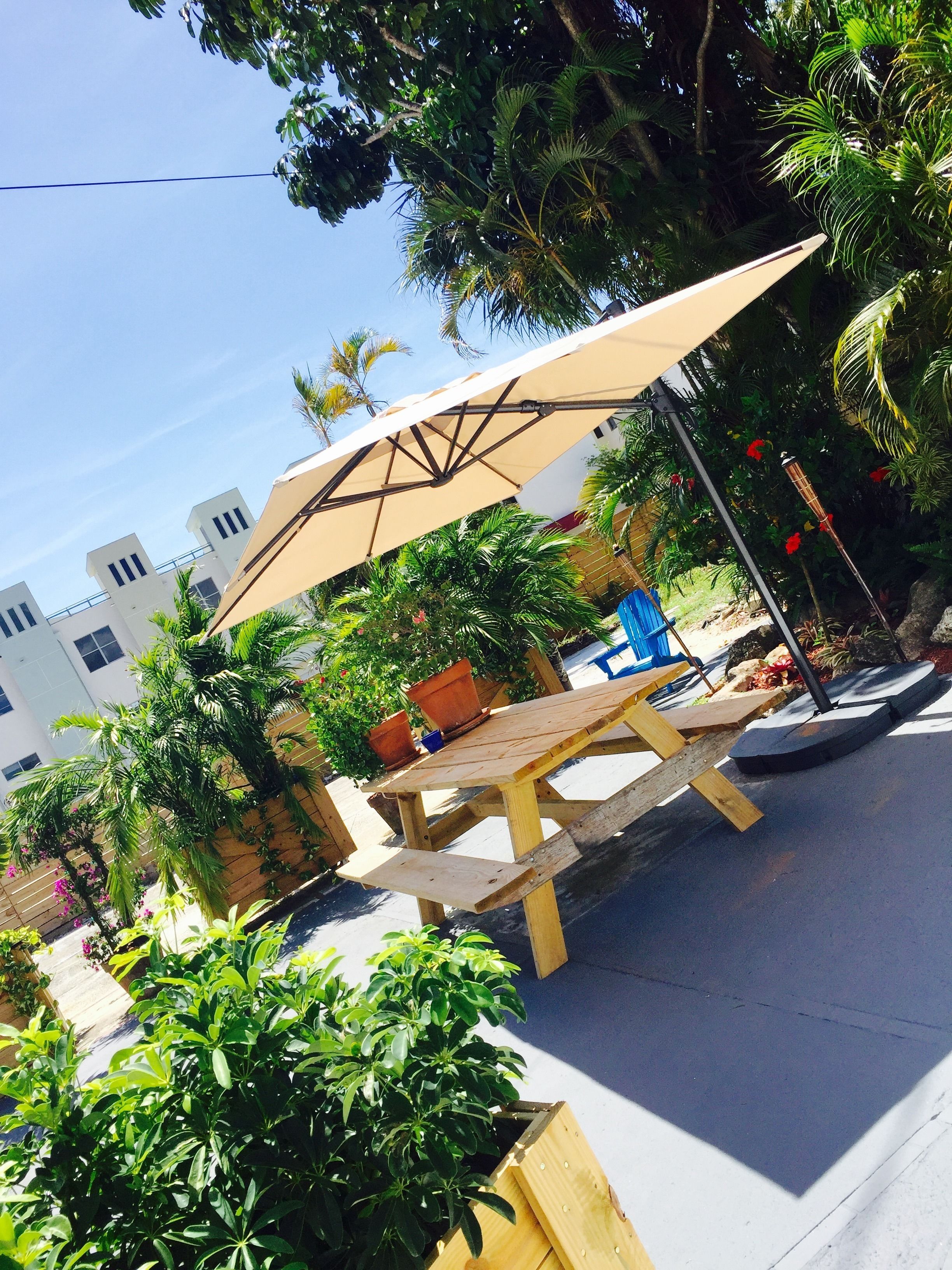 An outdoor seating area with a wooden table and a large umbrella, surrounded by lush greenery and a building in the background.