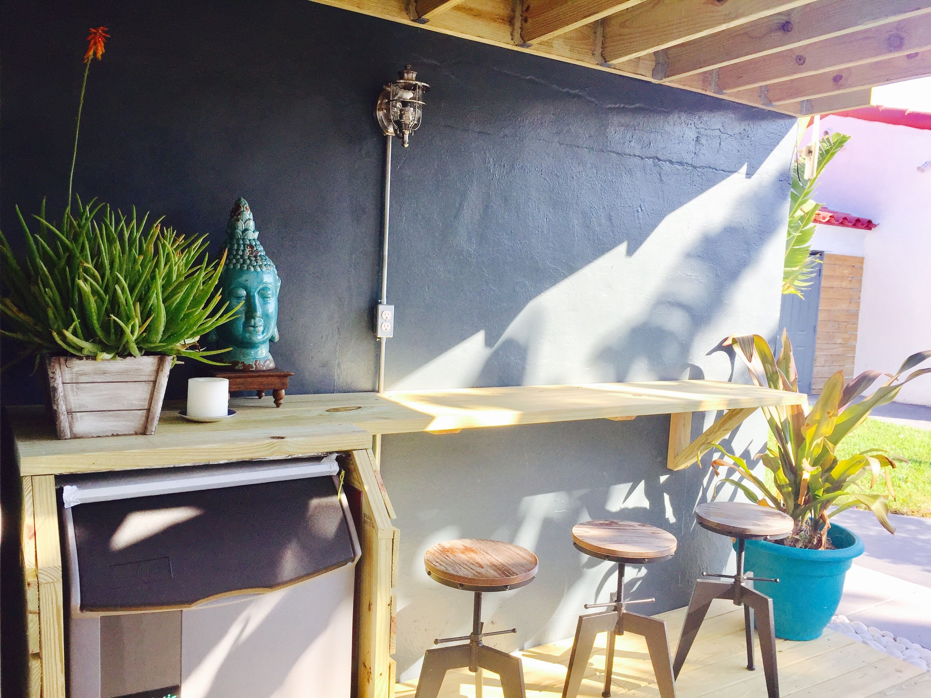 A wooden outdoor bar area with a blue wall, a Buddha statue, and potted plants.