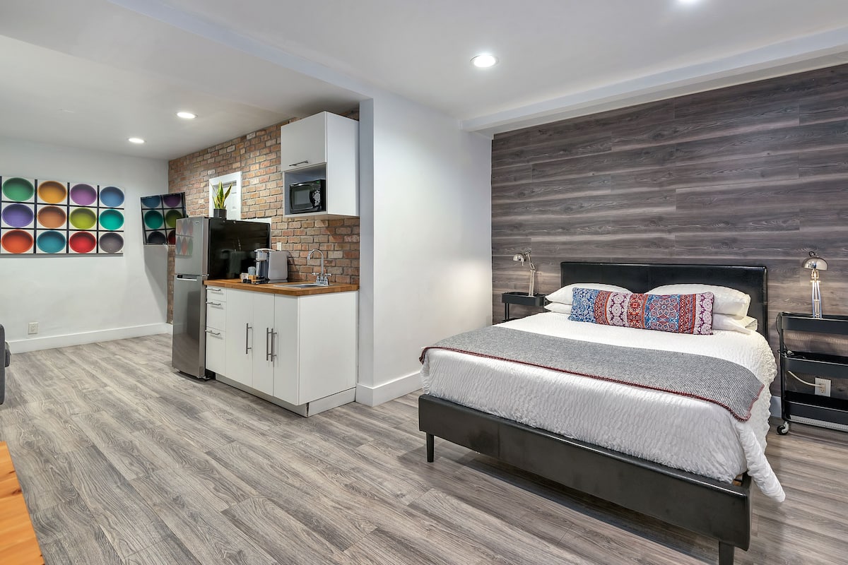 A modern bedroom with a large bed, a built-in TV, a kitchen area with a microwave, and a brick accent wall.