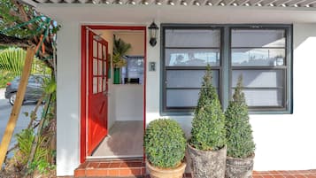 Una entrada de edificio con una puerta roja, un ventanal con persianas y tres macetas con plantas.