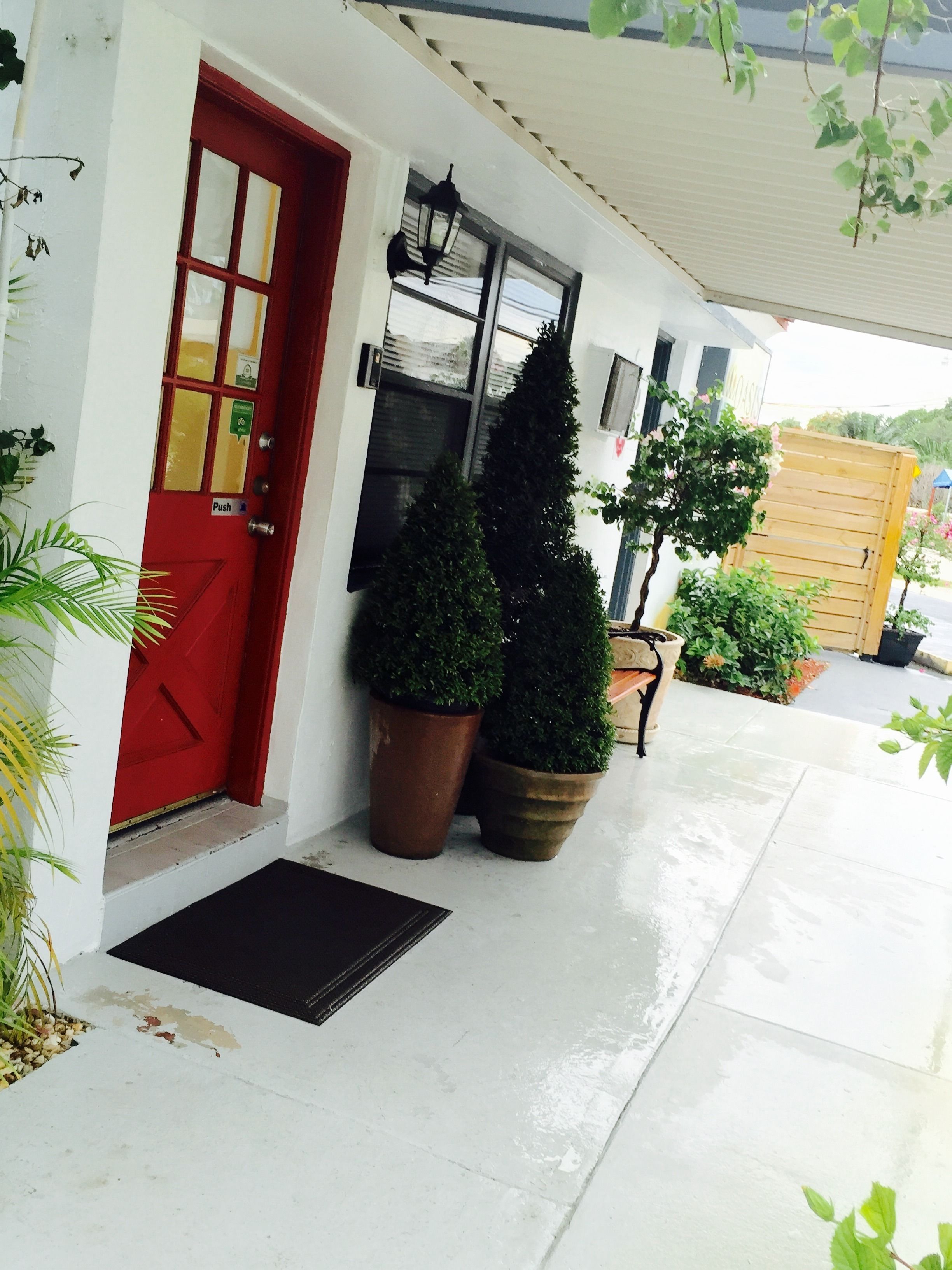 Una puerta roja con un botón pulsador, dos macetas con plantas, un banco y una valla de madera.