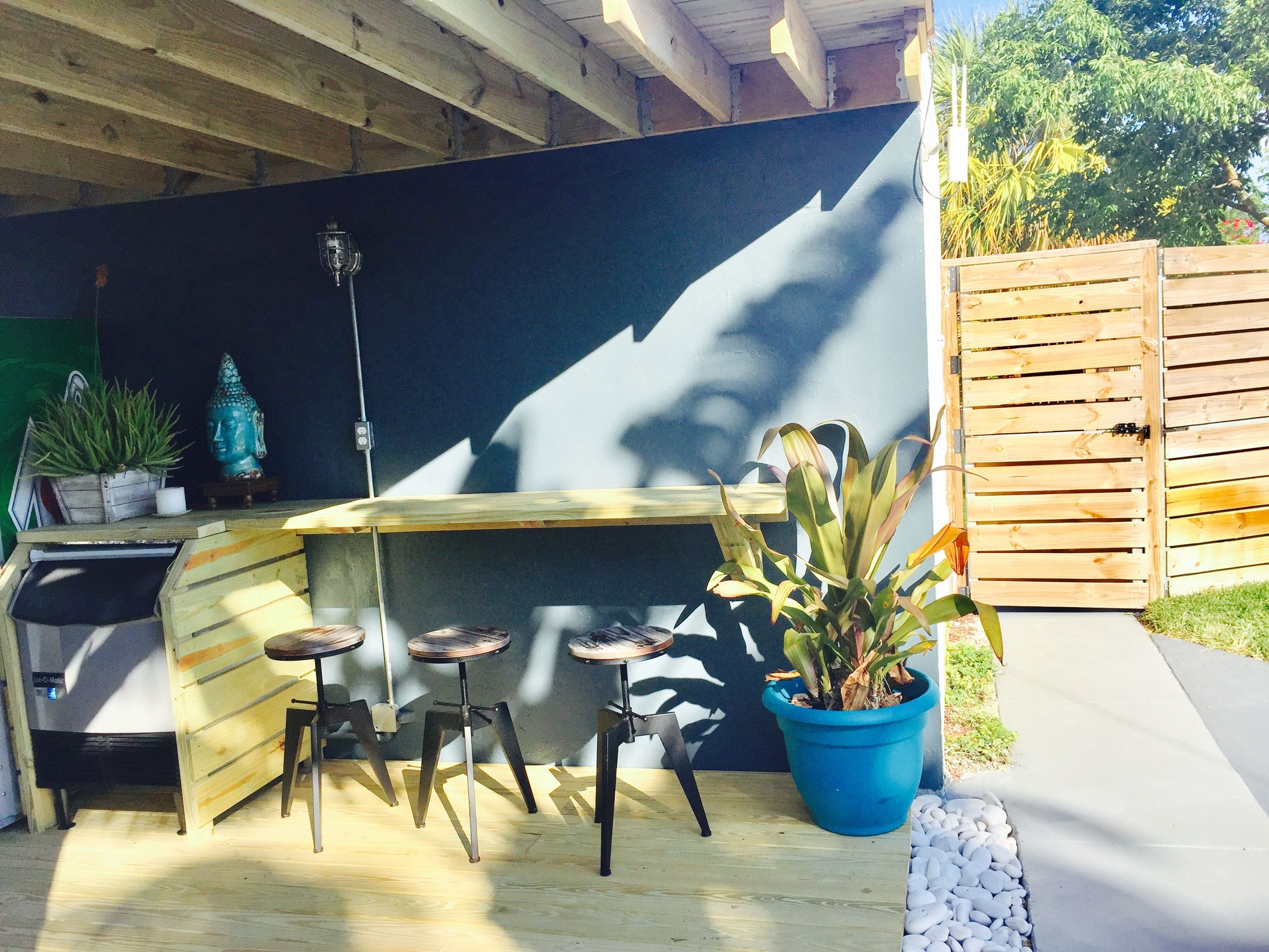 Eine Holzterrasse mit integrierter Bar, drei Barhocker, einem Blumentopf und einer Buddha-Statue.