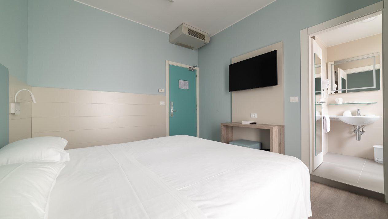 A hotel room with a bed, a television mounted on the wall, a bathroom with a sink and mirror, and a door leading to another room.