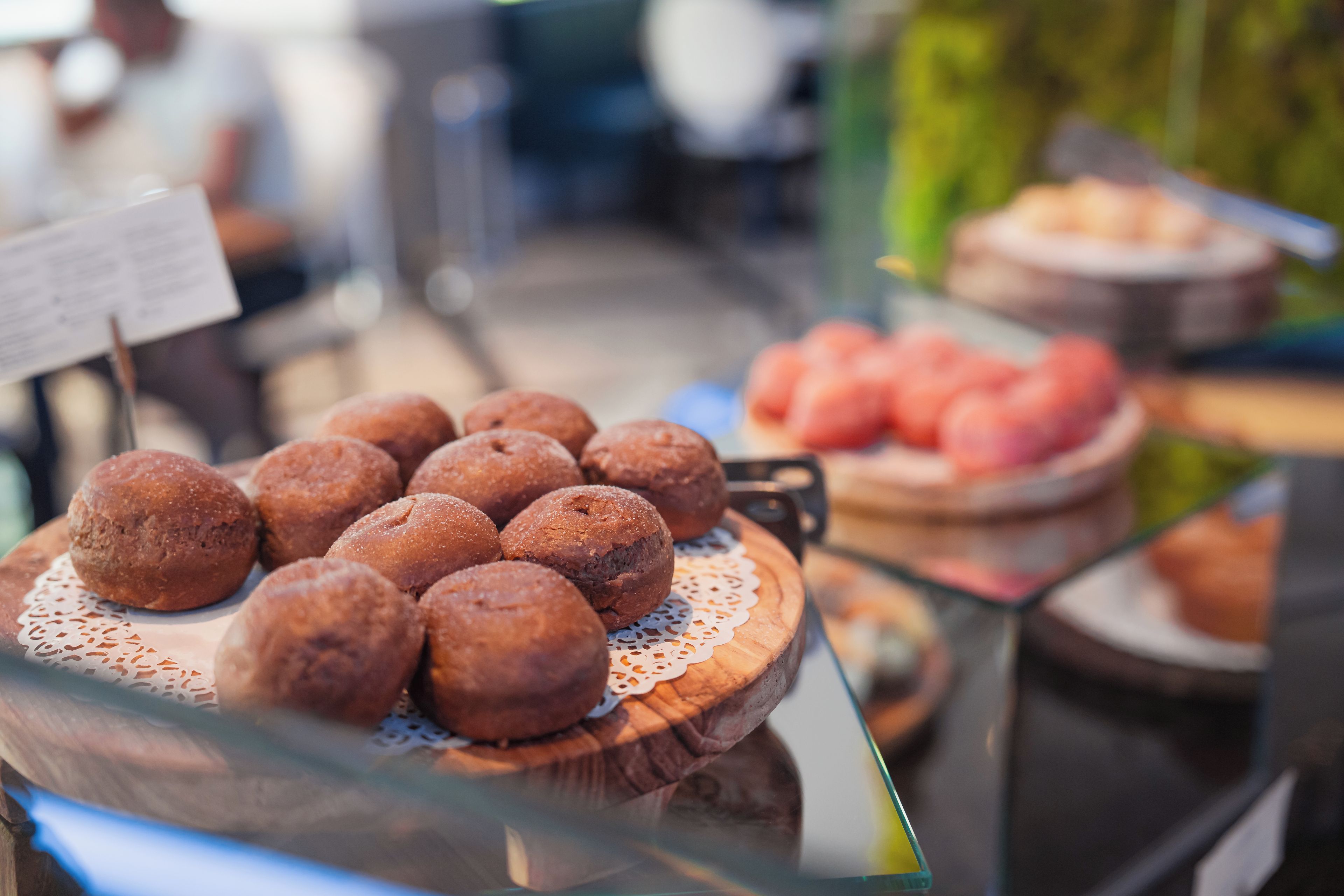 A display of round pastries on a wooden board with a lace doily, and a blurred background suggesting a public setting.