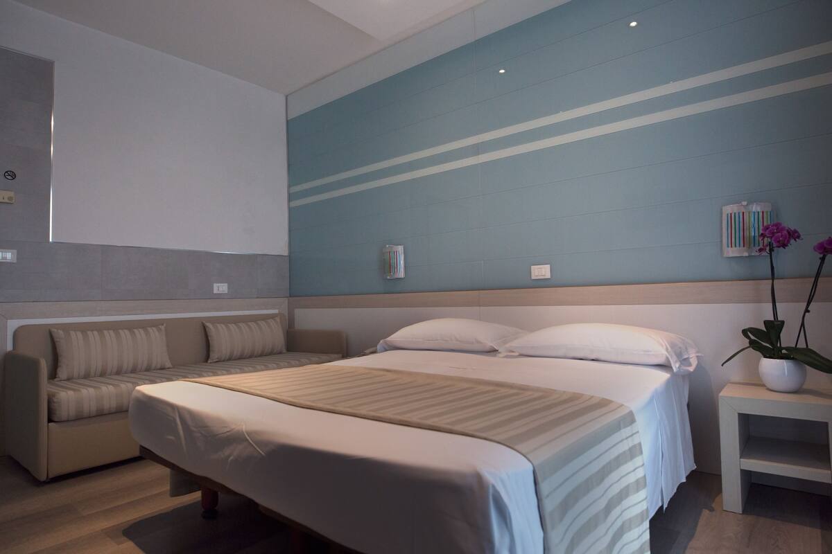 A modern hotel room with a large bed, a sofa, a small table with a plant, and a wall with a blue stripe design.