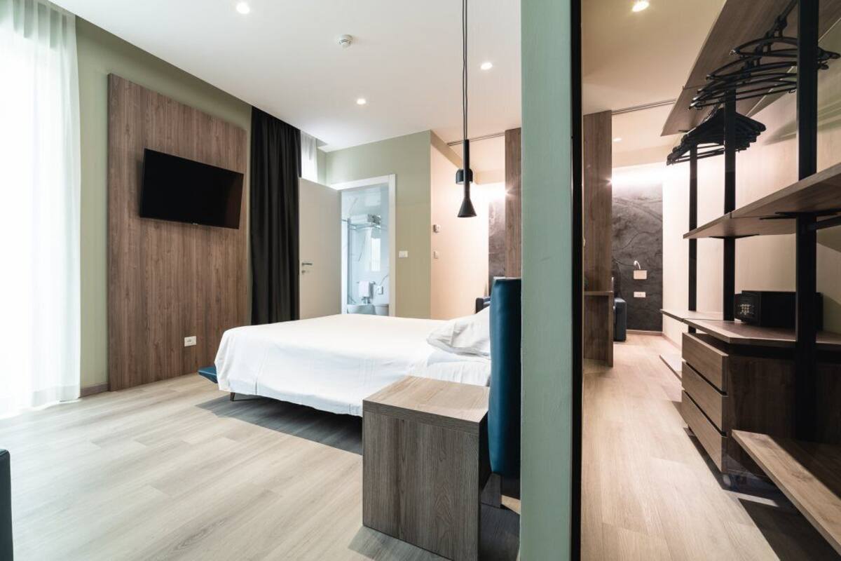 A modern hotel room with a large bed, a TV mounted on the wall, a wooden dresser, and a walk-in closet.