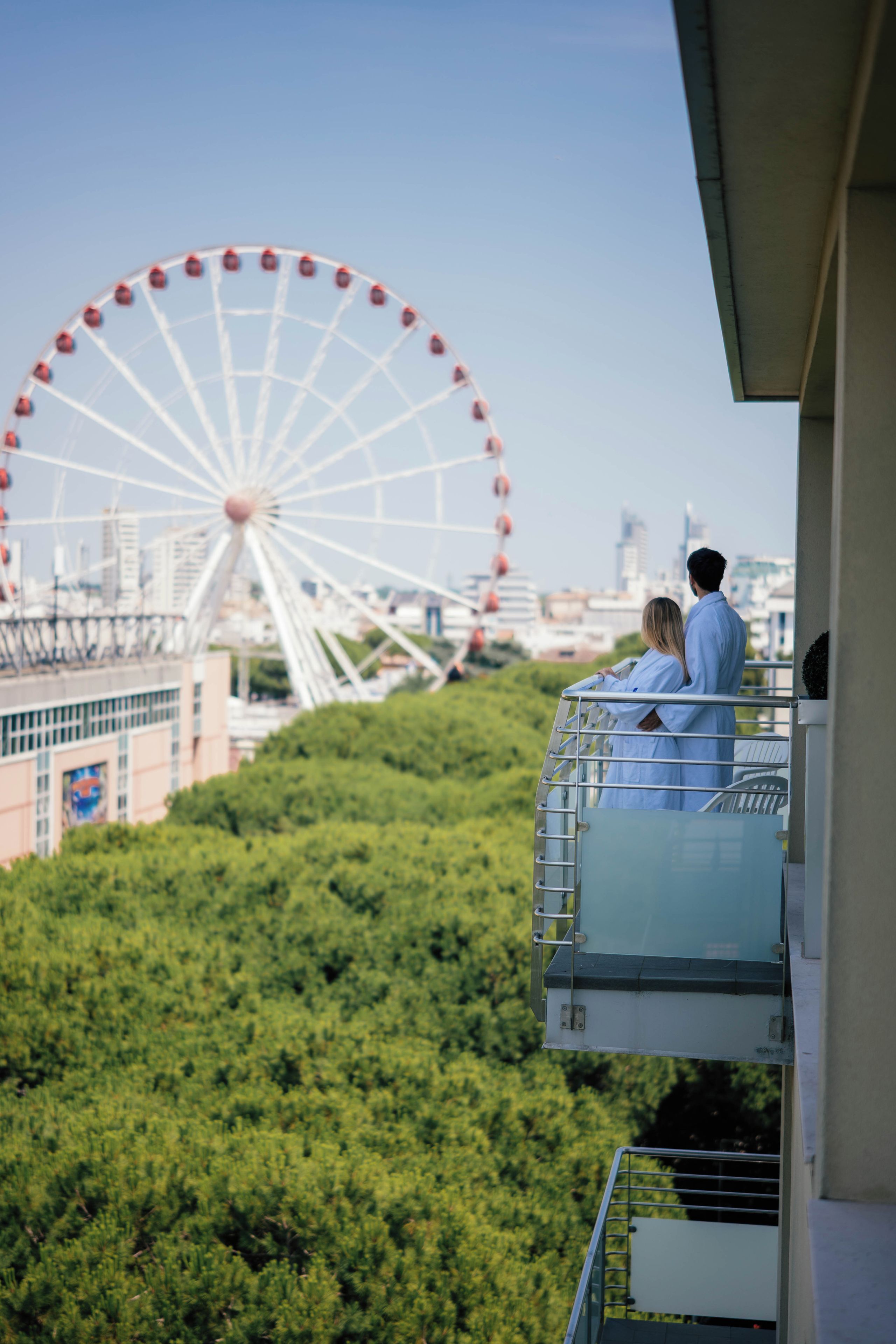 Una pareja en un balcón con vista a un paisaje urbano, con una noria grande al fondo.