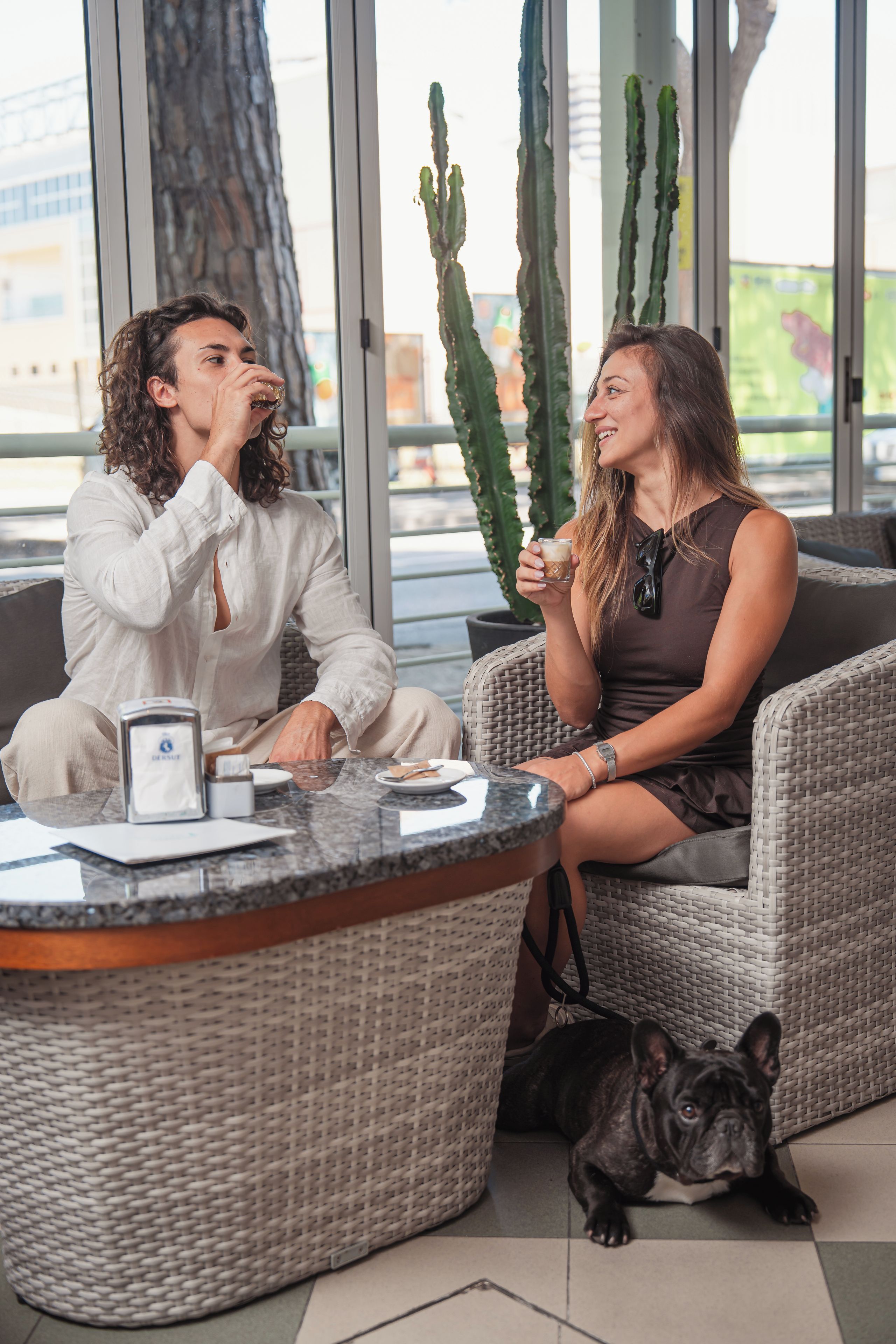 Zwei Personen sitzen an einem runden Tisch mit einer Kaffeemaschine, ein Hund liegt auf dem Boden und im Hintergrund sind große Kakteen zu sehen.
