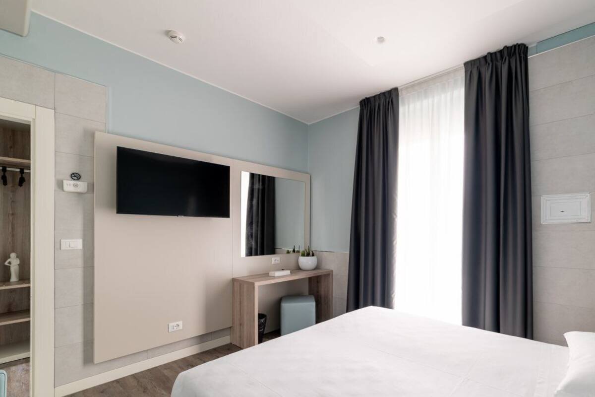 A modern hotel room with a large bed, a flat-screen TV mounted on the wall, a desk with a chair, and a wardrobe.