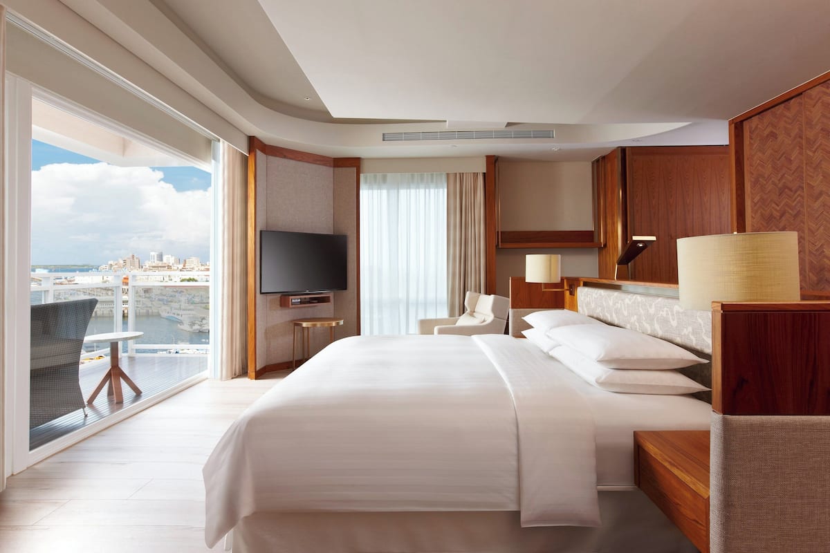 Habitación de hotel con una cama grande, televisor, silla, escritorio y vistas a la ciudad a través de un ventanal.