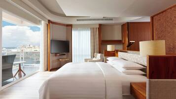 Een hotelkamer met een groot bed, een televisie, een zithoek met een stoel en een tafel, en uitzicht op de stad door het raam, met een houten hoofdeinde.